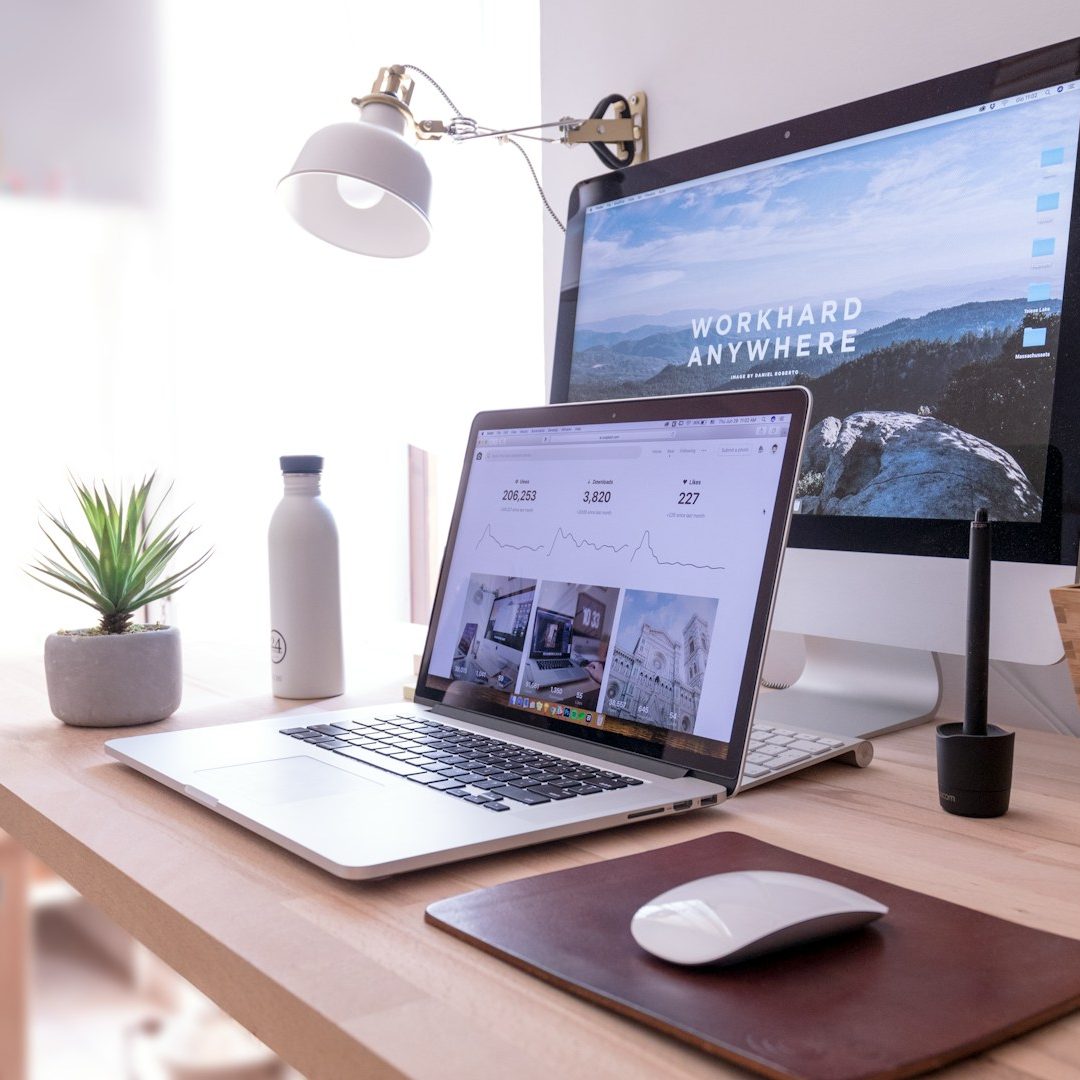 A modern workspace featuring a laptop, desktop monitor, plant, and water bottle.
